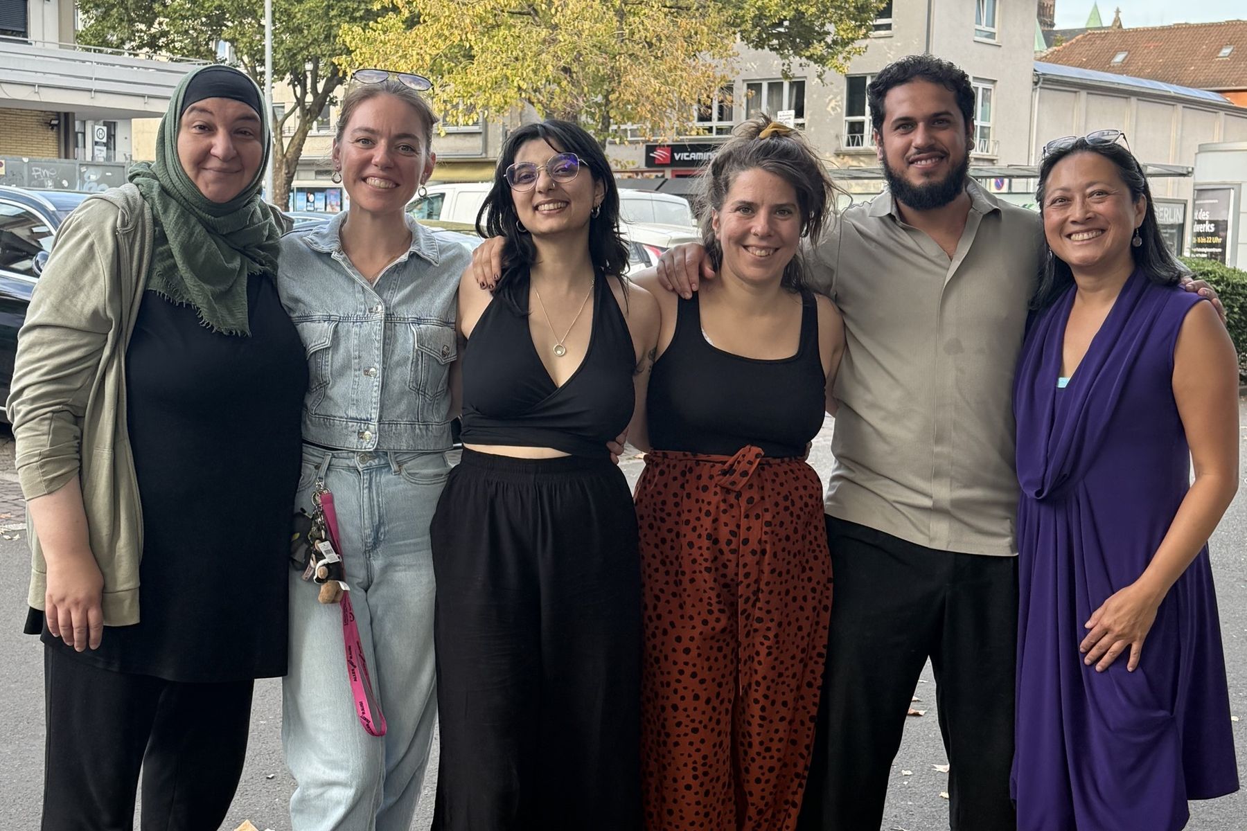 Gruppenfoto: sechs Menschen stehen lächelnd Arm in Arm auf einem Platz in der Stadt